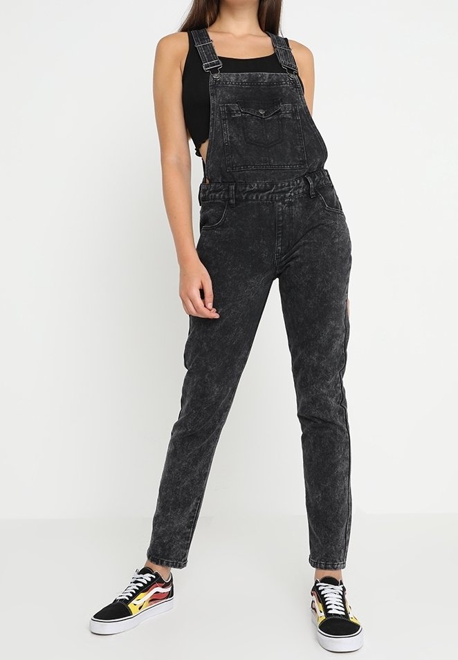 Femme portant une salopette en jean noir délavé à l'acide, un haut noir sans manches et des baskets noires avec des motifs de flammes, debout devant un fond blanc uni.