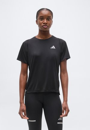 Mujer con camiseta negra de manga corta y leggings Adidas, de pie de frente contra un fondo liso.