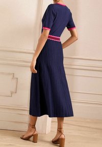 Robe en tricot côtelé marine avec des accents festonnés roses le long de l'encolure et de la taille ; présente une jupe évasée de longueur mi-mollet et des sandales à talons carrés.