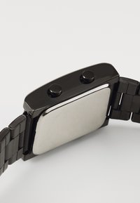 Montre en métal noir avec un boîtier rectangulaire, deux boutons poussoirs et un bracelet métallique composé de maillons entrelacés.
