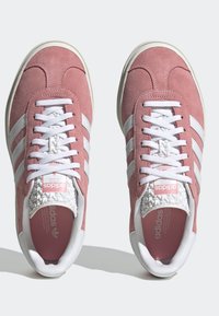 Un par de zapatillas Adidas de ante rosa con rayas blancas, cordones blancos y detalle texturizado en la lengua blanca, vistas desde arriba.