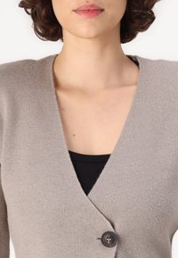 Beige cardigan med dyp V-hals, strikket tekstur og svart knappelukking i livet. Båret over en svart topp.