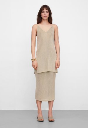 Kobieta o brązowych włosach, ubrana w beżową, bezrękawnikową, warstwową dzianinową sukienkę z dekoltem w serek, beżowe sandały na obcasie z odkrytymi palcami i złote bransoletki, stoi na tle białej ściany.