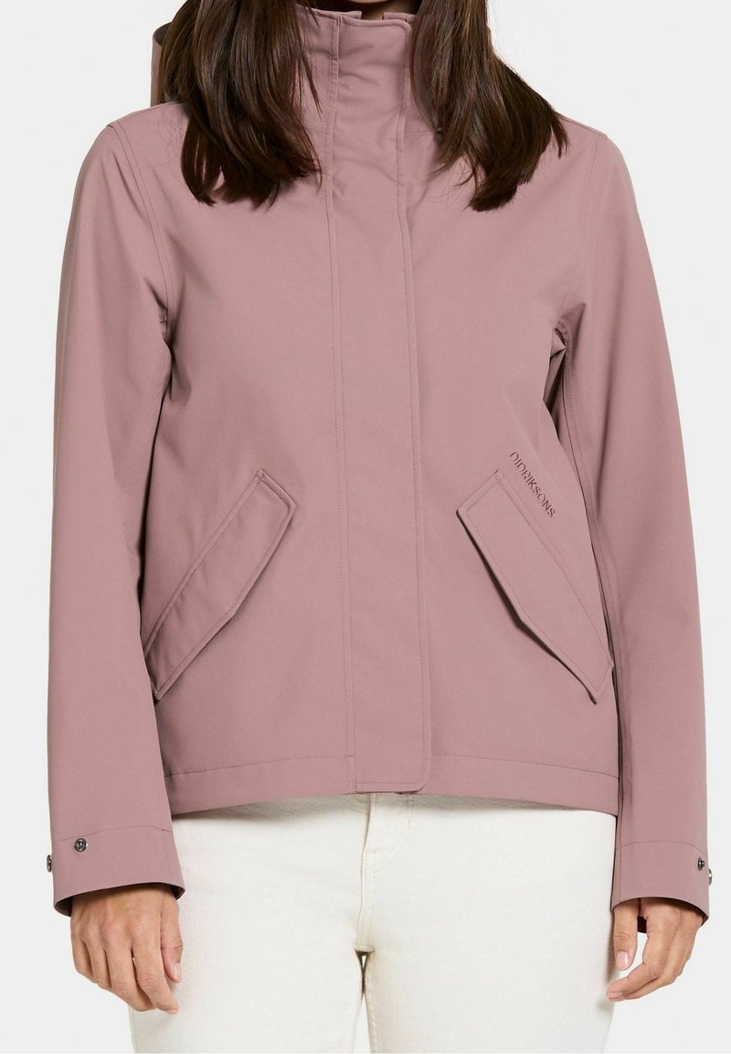 Femme portant une veste imperméable rose poussière avec des poches à rabat angulaires et un col haut, associée à un pantalon blanc cassé.