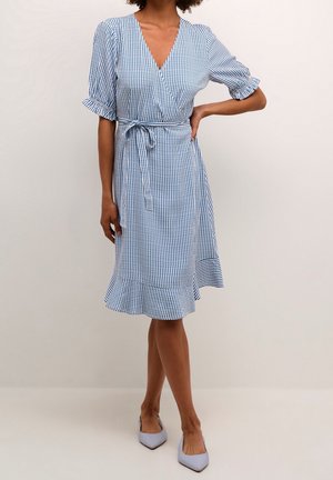 Donna che indossa un vestito a portafoglio a righe verticali blu e bianco con maniche corte a sbuffo, cintura annodata in vita e orlo arricciato, in piedi con una mano sul fianco.
