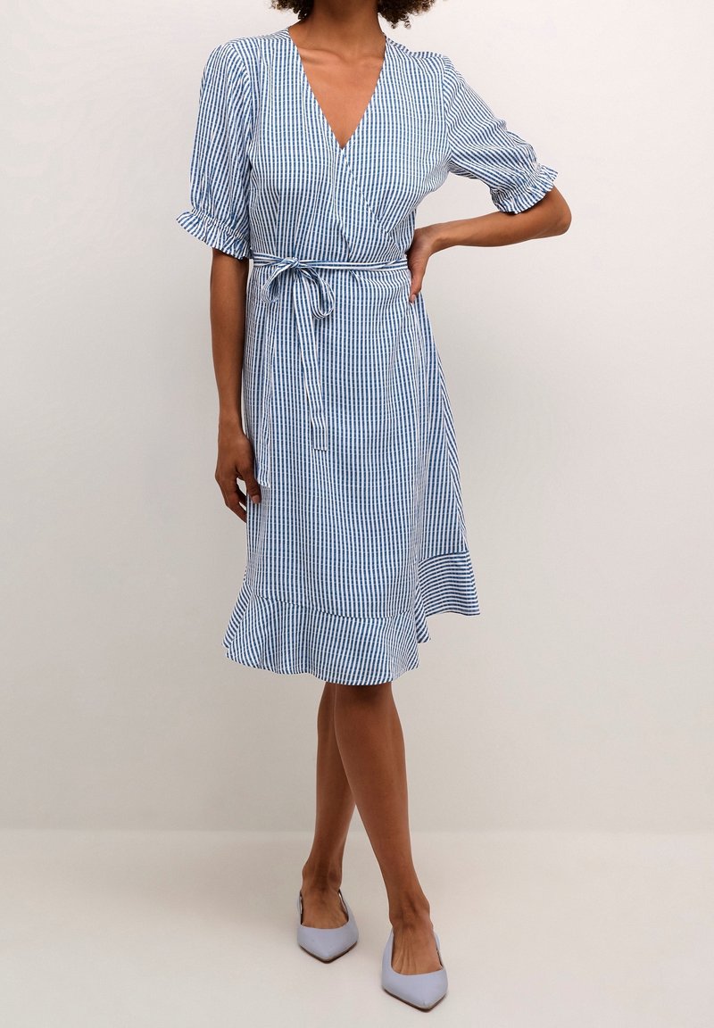 Femme portant une robe portefeuille rayée verticalement bleu et blanc avec manches courtes bouffantes, ceinture nouée à la taille et ourlet à volants, debout avec une main sur la hanche.