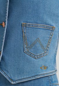 Υφασμα από ανοιχτό μπλε τζιν με ραμμένη πίσω τσέπη που διαθέτει πορτοκαλί ραφή και ένα μικρό κεντημένο "LW" κοντά στην κάτω άκρη.