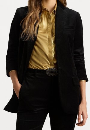 Kvinde iført sort fløjlsblazer og bukser med en gylden satin skjorte stukket ind, hænderne i lommerne og en dekorativ bæltespænde.
