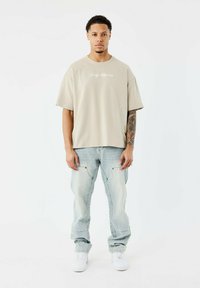 Camiseta beige de gran tamaño con bordado de cactus y jeans baggy azul claro. El modelo lleva zapatillas blancas, presentando una silueta relajada.
