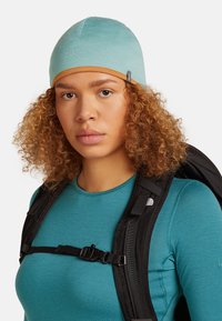 Young person with curly hair wearing a teal beanie, matching top, and black backpack with chest strap against a white background.