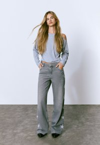 Young woman with long blonde hair wearing a gray cold-shoulder top, wide-leg gray jeans with star patterns, and black shoes, standing indoors.