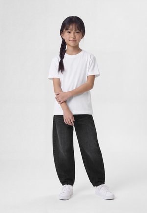 Niño con el pelo trenzado, lleva una camiseta blanca, pantalones negros holgados y zapatillas blancas, de pie con los brazos cruzados delante de un fondo blanco liso.