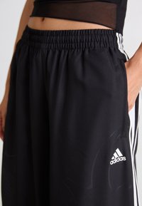 Mustat urheiluhousut, jotka on valmistettu sileästä kankaasta ja joissa on joustava vyötärö, valkoinen Adidas-logo ja valkoiset sivuraidat.