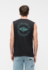Άνδρας που φοράει μαύρο αμάνικο μπλουζάκι Rip Curl Surf Co και ανοιχτόχρωμο μπλε τζιν, στέκεται γυρισμένος ανάποδα σε απλό φόντο.