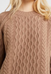Jasnobrązowy sweter dzianinowy z teksturowanym wzorem splotu warkoczowego oraz ściągaczem przy dekolcie, prezentujący miękkie włókna i gruby fason.