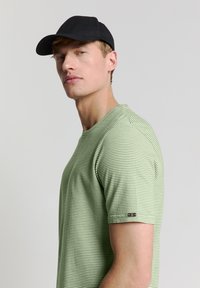 Groen gestreept T-shirt van zacht materiaal, met korte mouwen en een ronde hals. Gedragen met een zwarte pet; subtiel logo op de mouw.