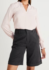 Ljusrosa långärmad topp med V-ringning och samlade manschetter, tillsammans med mörkgrå randiga shorts med rak passform och uppvikt nederkant.