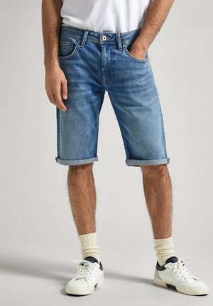 Shorts de mezclilla azul claro con dobladillos enrollados, cinco bolsillos y costuras naranjas. Combinados con zapatillas blancas y calcetines crema.