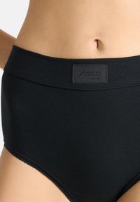 Svarta högmidjade trosor tillverkade av mjukt, stretchigt material. Har en slät midjeband med logotyp och vågiga kanter vid benöppningarna.