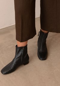 Persona che indossa pantaloni marroni corti e stivaletti neri in pelle con cerniere laterali, in piedi su un pavimento beige.