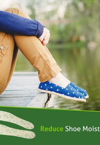 Blue espadrilles with white polka dots and a jute sole. Tan trousers and a navy sweater are worn, seated on a wooden dock by water.