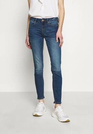 Jeans skinny de mezclilla azul con cintura de altura media, cinco bolsillos y costuras en contraste. Llevado con zapatillas blancas y una blusa blanca.