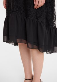 Schwarzes Kleid mit aufwendiger Spitze am oberen Teil, geschichtet mit einem durchsichtigen, rüschierten Saum. Das Gesamtdesign ist elegant und fließend.