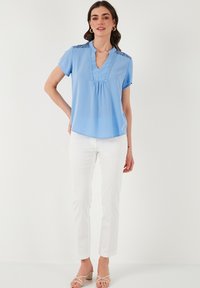 Blusa azzurra chiara a maniche corte con scollatura a V, cuciture decorative e dettagli in pizzo sulle spalle, abbinata a pantaloni bianchi slim e sandali beige.