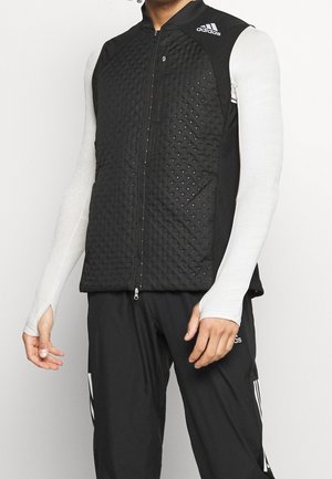 Mężczyzna ubrany w czarną pikowaną kamizelkę bez rękawów Adidas na białej koszuli z długim rękawem i czarne spodnie dresowe Adidas, stoi na białym tle.