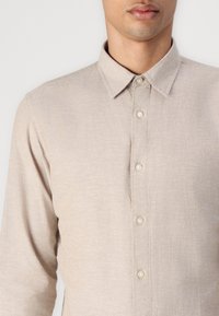 Beige, long-sleeve shirt with a button-down collar, featuring a subtle texture and visible stitching; five buttons down the front.