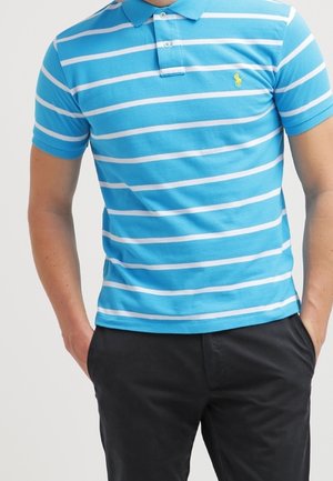 Homme portant un polo à manches courtes rayé bleu et blanc avec un pantalon noir, les mains dans les poches, devant un fond uni.