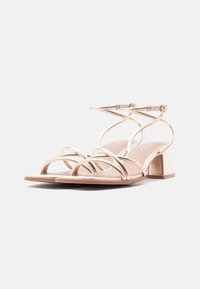 Ζευγάρι από χρυσά σανδάλια με λουριά και χοντρό τακούνι, με αγκράφες στον αστράγαλο, σε απλό λευκό φόντο.