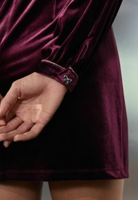 Violettes Samtkleid mit langen Ärmeln, das mit einem Umschlag mit einer kleinen Schleifenakzent und dekorativen Perlen entlang des Randes ausgestattet ist. Glatte Textur.