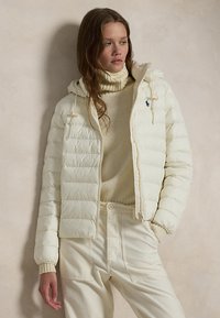 Frau trägt cremefarbene gesteppte Kapuzenjacke über beigem Rollkragenpullover und cremefarbenen hoch taillierten Hosen, steht vor neutralem Hintergrund.