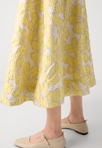 Jupe maxi à motifs floraux jaunes en tissu texturé, avec un ourlet élégant. Des ballerines beiges avec une lanière complètent la tenue.