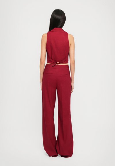 Bordeaux-farbenes ärmelloses Crop-Top mit hohem Kragen, der am Rücken gebunden wird. Weit geschnittene Hose in passender Farbe mit Seitentaschen und glatter Textur.