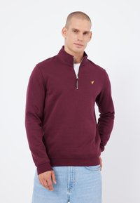 Burgundská mikina s čtvrtovým zipem a žebrovaným límcem, přední zip a malé žluté logo na hrudi. Měkká, texturovaná látka.
