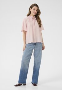 Blouse en coton rose avec un col volanté et des liens, associée à un jean à jambes larges bleu clair et des mules à talons bruns, présentée sur une silhouette féminine.