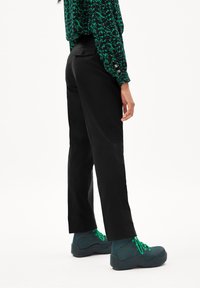 Černé kalhoty se střihem na míru, s jemným leskem, v kombinaci se zelenými šněrovacími botami. Součástí outfitu je vzorovaná blůza.