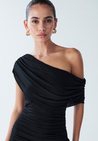 Robe noire épaules dénudées en tissu lisse avec des détails drapés et une texture froncée ; complétée par des boucles d'oreilles dorées imposantes.