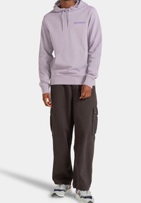 Persona che indossa un felpa leggera viola con la scritta "ELEMENT", pantaloni cargo neri e sneakers bianche con dettagli blu e rossi.