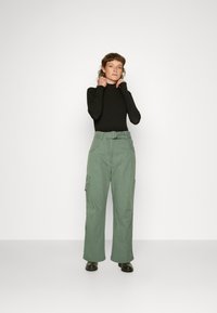 Top de gola alta preto combinado com calças cargo largas verdes, com bolsos laterais e um cinto, em um fundo claro.