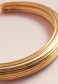 Gul armring med en glat, rundet design med flere riller, der skaber et struktureret mønster. Overfladen har en poleret finish.