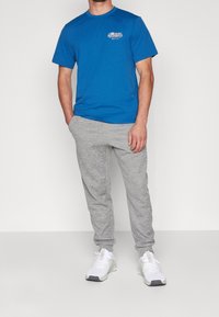 Camiseta de manga corta azul con logo en el pecho, combinada con pantalones de chándal gris claro y zapatillas deportivas blancas. Look deportivo casual.
