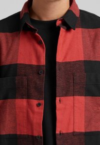 La camicia in flanella a quadri rossi e neri presenta un frontale con bottoni, due tasche sul petto e un colletto. Il tessuto appare morbido e strutturato.