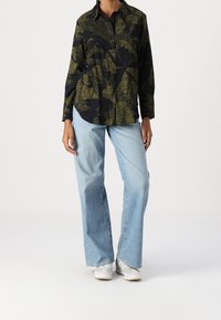 Camicia nera con pattern astratto verde, stile button-up, maniche lunghe, abbinata a jeans a gamba larga blu chiaro e sneakers grigie.