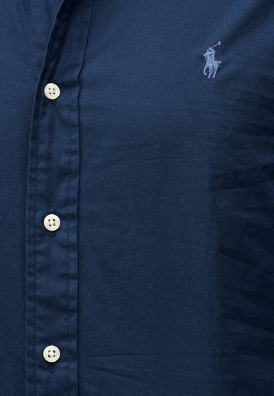 Tmavomodrá košeľa na gombíky s bielymi gombíkmi a malým vyšívaným logom hráča póla na ľavej strane hrude.