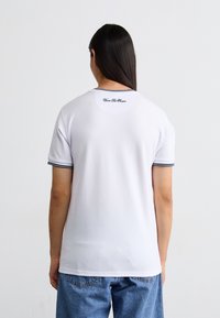 Hvid t-shirt med korte ærmer og rund halsudskæring, forsynet med kontrast i marineblå og logo-detaljer på den øverste del af ryggen. Blødt, tekstureret stof.