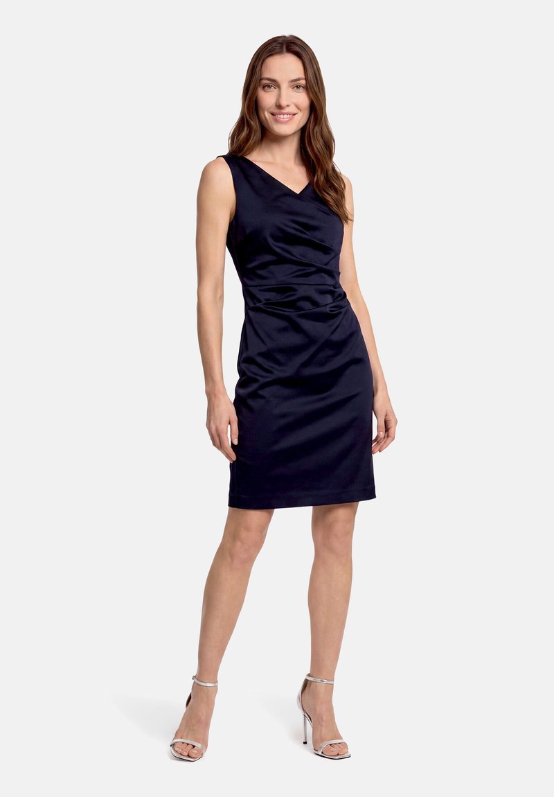 Femme debout portant une robe bleu marine sans manches longueur genoux avec des sandales argentées à talons hauts sur fond blanc.