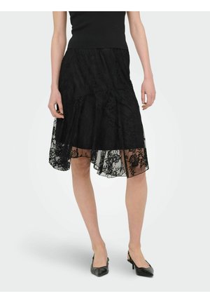 Žena nosí čiernu čipkovanú sukňu do kolien s asymetrickým lemom a čierne lodičky s ostrou špičkou na bielom pozadí.